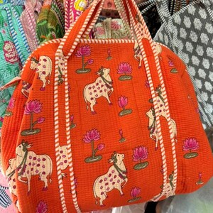 May include: A quilted orange tote bag featuring a repeating pattern of white cows with pink spots and pink lotus flowers. The bag has two matching orange and white striped fabric handles and a quilted texture throughout the entire exterior surface.