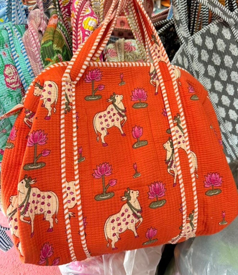 May include: A quilted orange tote bag featuring a repeating pattern of white cows with pink spots and pink lotus flowers. The bag has two matching orange and white striped fabric handles and a quilted texture throughout the entire exterior surface.