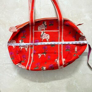 May include: A bright orange quilted tote bag featuring a repeating pattern of white cows and pink lotus flowers. The bag has two striped handles and a zipper closure. A white measuring tape across the bag shows a width of approximately 56 centimetres.