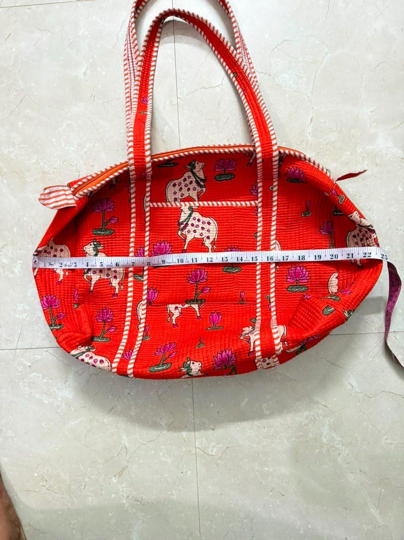 May include: A bright orange quilted tote bag featuring a repeating pattern of white cows and pink lotus flowers. The bag has two striped handles and a zipper closure. A white measuring tape across the bag shows a width of approximately 56 centimetres.