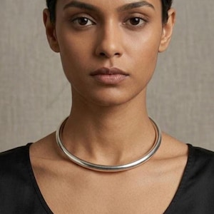 Puede incluir: Un collar tipo gargantilla de metal plateado, liso y pulido, con un diseño tubular redondeado. El collar se ajusta a la base del cuello, presentando una banda continua y sin costuras con una superficie reflectante que capta la luz.
