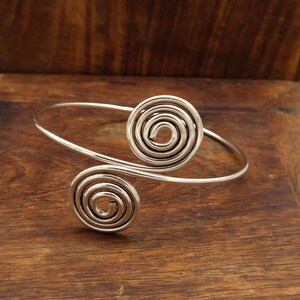 May include: A silver-toned open cuff bracelet featuring two prominent spiral motifs at each end. The metal band is smooth and circular, with the two coils positioned to face opposite directions, creating a balanced and symmetrical design.