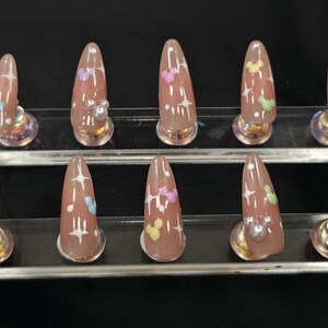 May include: Set of ten almond-shaped, press-on nails. Each nail is a nude color with white starburst designs and pastel-colored butterfly and Mickey Mouse-shaped accents. The nails are displayed on a clear stand against a black background.