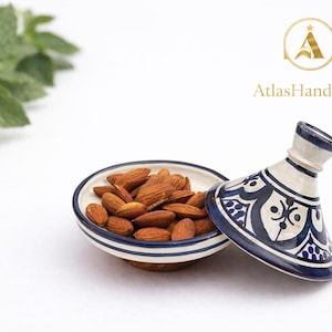Può includere: Un piatto di ceramica pieno di mandorle, con un coperchio abbinato. Il piatto e il coperchio sono bianchi con motivi geometrici blu. Il testo "AtlasHandmade" è in oro. Foglie di menta fresca sullo sfondo.