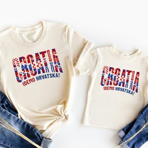 two white t-shirts with the word "Croatia" printed on them, along with a smaller child's t-shirt. The t-shirts are laid out on a white background, and there are also some jeans visible in the image.