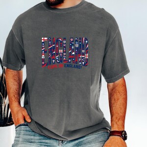 A man wearing a grey t-shirt with the word "ENGLAND" printed on it, along with various symbols and text related to the United Kingdom.