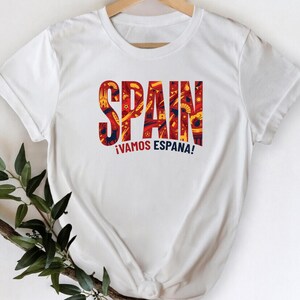 Può includere: T-shirt bianca con la parola "SPAIN" a grandi lettere colorate. Le lettere sono riempite con un motivo di disegni rossi, gialli e blu. Sotto la parola "SPAIN" c'è la frase "¡VAMOS ESPAÑA!"