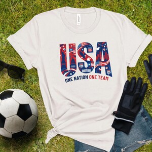 a white t-shirt with the text "USA" printed on it, along with a soccer ball and some gloves, all laid out on a grassy surface.