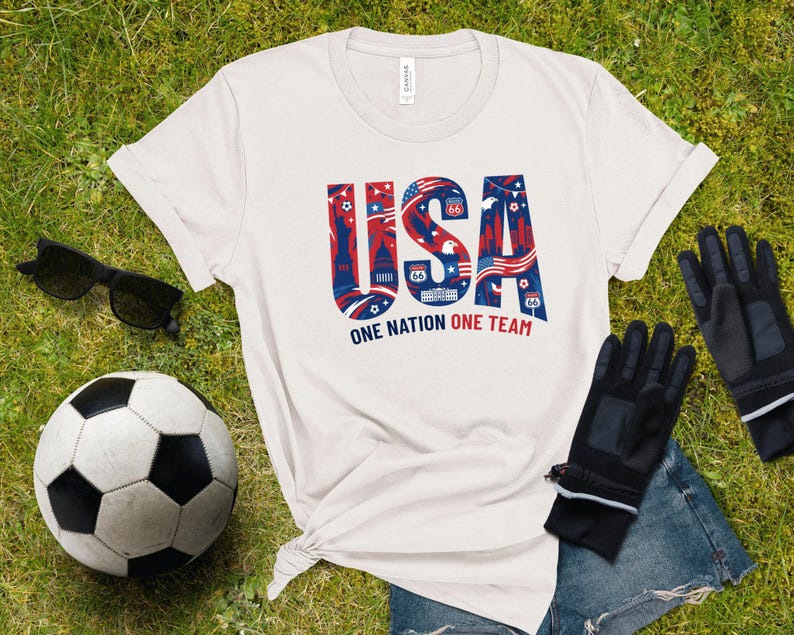 a white t-shirt with the text "USA" printed on it, along with a soccer ball and some gloves, all laid out on a grassy surface.