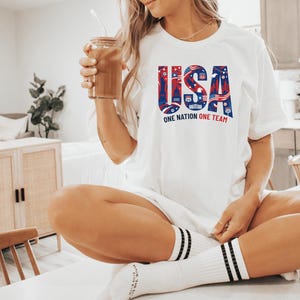 A woman wearing a white t-shirt with the text "USA" and "ONE NATION ONE TEAM" is sitting on a table, holding a drink in her hand.