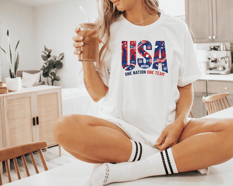 A woman wearing a white t-shirt with the text "USA" and "ONE NATION ONE TEAM" is sitting on a table, holding a drink in her hand.