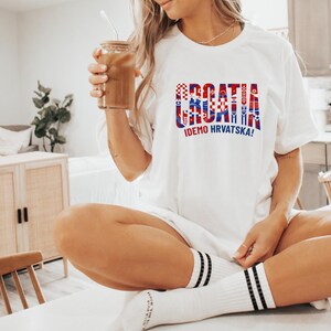 A woman wearing a white t-shirt with the word "Croatia" printed on it, sitting on a chair in a kitchen. She is holding a drink in her hand.