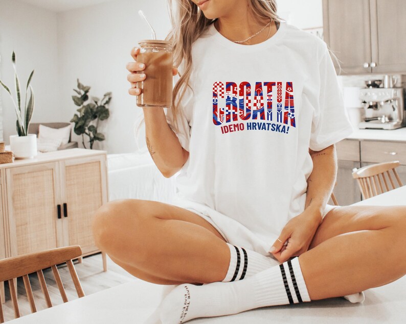 A woman wearing a white t-shirt with the word "Croatia" printed on it, sitting on a chair in a kitchen. She is holding a drink in her hand.