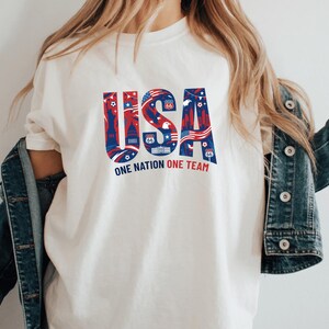 A person wearing a white t-shirt with the text "USA" and "ONE NATION ONE TEAM" printed on it, along with various patriotic symbols.