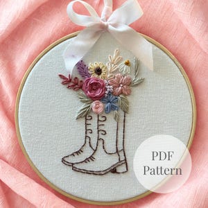 Puede incluir: Un bastidor de bordado circular de madera que contiene tela blanca con un diseño cosido a mano de dos botas llenas de un colorido ramo de flores. Una cinta blanca está atada en la parte superior del bastidor. El texto PDF Pattern es visible.