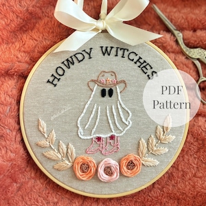 Puede incluir: Un bastidor de bordado circular con tela beige y el texto bordado HOWDY WITCHES. El diseño muestra un fantasma blanco con sombrero de vaquero marrón y botas rosas, rodeado de flores y hojas bordadas en tonos naranja y rosa.