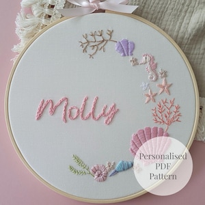 Puede incluir: Un bastidor de bordado circular de madera que contiene tela blanca con el nombre Molly bordado en hilo rosa. El diseño presenta una corona de elementos marinos bordados que incluyen un caballito de mar, coral y varias conchas en tonos pastel.