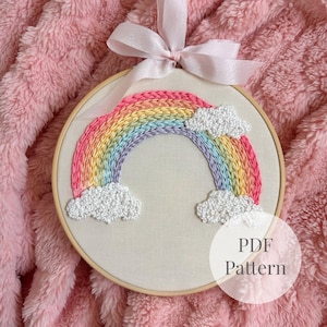 Puede incluir: Un bastidor de bordado circular de madera con un diseño de arcoíris cosido a mano sobre tela color hueso. El arcoíris presenta bandas de hilo rosa, naranja, amarillo, verde y azul con nubes blancas texturizadas. Un lazo rosa claro está atado arriba.