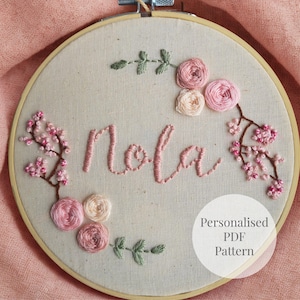 Puede incluir: Aro de bordado con el nombre "Nola" en hilo rosa. El diseño incluye rosas rosas y crema, ramas de flores de cerezo y hojas verdes. También se incluye el texto "Personalised PDF Pattern".