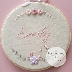 Puede incluir: Arte de aro bordado con el nombre "Emily" en escritura rosa. El diseño incluye flores bordadas, hojas y mariposas en tonos rosa y lavanda, con un aro de madera y una cinta.