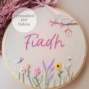Puede incluir: Un bastidor de bordado con el nombre Fiadh cosido en hilo rosa. El diseño incluye dos pájaros en una rama con una casita, una mariposa azul, una abeja y varias flores coloridas como lavanda y girasoles sobre una tela de color beige.