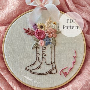 Puede incluir: Arte de aro bordado con un par de botas de vaquero marrones llenas de flores coloridas. Las flores incluyen rosas rosas, margaritas y flores azules y melocotón. El texto "PDF Pattern" está en un círculo blanco. Una cinta rosa está atada en la parte superior.