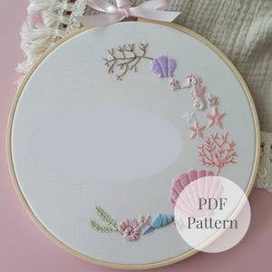 Puede incluir: Un bastidor de bordado circular de madera que contiene tela blanca con un diseño bordado a mano de vida marina, incluyendo un caballito de mar, coral y varias conchas en tonos pastel de rosa, morado, azul y verde, con una cinta rosa.