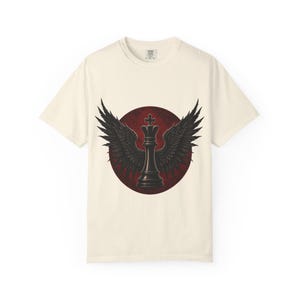 Könnte beinhalten: Cremefarbenes T-Shirt mit einem schwarzen Schachkönig mit gefiederten Flügeln, vor einem dunkelroten kreisförmigen Hintergrund. Das grafische Design ist mittig platziert. Das T-Shirt hat kurze Ärmel und einen Rundhalsausschnitt.