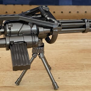 Könnte beinhalten: Ein metallisch dunkelgraues Spielzeug-Minigun mit rotierender Laufbaugruppe, einem oben montierten Griff, einem rechteckigen Munitionsmagazin und einem Stativ. Das Waffenmodell hat eine glänzende Oberfläche und besteht aus vielen Teilen.