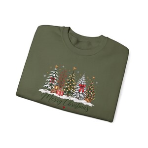 Könnte beinhalten: Ein olivgrünes Sweatshirt mit Rundhalsausschnitt und einem festlichen Motiv aus vier geschmückten Weihnachtsbäumen, einer Laterne und verpackten Geschenken auf einer Schneefläche. Der Schriftzug Merry Christmas steht in dunkelgrüner Schreibschrift.