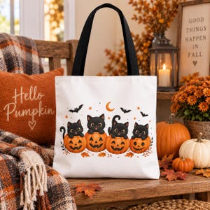 Könnte beinhalten: Eine weiße Tragetasche mit schwarzen Henkeln und einem Grafikdruck von vier schwarzen Kätzchen, die in orangefarbenen Halloween-Kürbissen sitzen, verziert mit kleinen Fledermäusen, Sternen und einer Mondsichel auf weißem Grund.