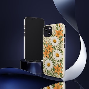 Könnte beinhalten: Eine Smartphone-Hülle mit einem botanischen Muster aus weißen Gänseblümchen und orangefarbenen Lilien mit grünem Blattwerk auf cremefarbenem Grund. Die Hülle hat einen schwarzen Kameraausschnitt und Seitentasten, neben einem Smartphone.