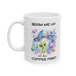 Könnte beinhalten: Ein weißer Keramikbecher mit einer bunten Weltraumgrafik, die einen grünen Außerirdischen zeigt, der unter einer fliegenden Untertasse ein Friedenszeichen macht. Der Text BEAM ME UP steht darüber und COFFEE FIRST steht darunter in schwarzer Schrift.