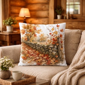 Peut inclure: Un coussin carré décoratif orné d'une illustration aquarelle imprimée représentant un paysage automnal. Le motif montre un muret en pierre bordé d'érables aux feuilles orange et rouges, jonchant le sol de teintes automnales.