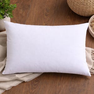 Peut inclure: Un oreiller rectangulaire blanc sur une surface en bois. L'oreiller est le point central, avec une apparence douce et moelleuse. Une couverture beige est partiellement visible, ainsi qu'une petite plante en pot et un plateau décoratif avec une bougie.
