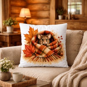 Peut inclure: Un coussin carré blanc orné d'une illustration centrale représentant une petite souris brune enveloppée dans une écharpe à carreaux orange et marron. Le motif comprend des feuilles d'érable automnales orange et rouges et un gland.