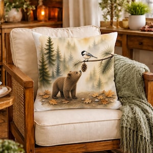May include: A square throw pillow featuring a watercolour-style illustration of a brown bear cub standing on a forest floor with fallen autumn leaves, looking up at a small black and white bird perched on a pine branch with a hanging pinecone.