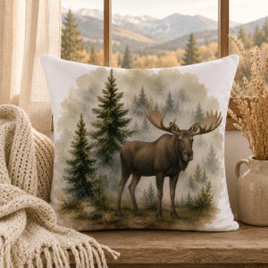 May include: A square throw pillow featuring a watercolour-style illustration of a brown moose standing in a forest of green pine trees. The background depicts a misty, soft-focus woodland scene with muted green and brown tones on a white fabric base.