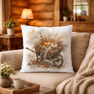Peut inclure: Un coussin carré blanc orné d'une illustration à l'aquarelle représentant une brouette en bois remplie de citrouilles orange et vertes, de blé séché et de feuilles d'automne, recouverte d'une couverture à carreaux sur un fond neutre.