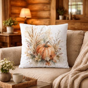May include: A square throw pillow featuring a watercolour illustration of an orange pumpkin surrounded by dried wheat stalks, autumn leaves, and small berries on a white background. The design uses soft, muted earth tones and artistic paint splatters.
