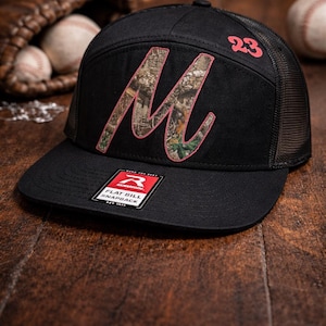 May include: Black snapback hat with a flat bill, featuring a large pink-outlined "M" with a camouflage pattern. The number "23" is on the side. A "Flat Bill Snapback" tag is visible. Baseball glove and balls are in the background.