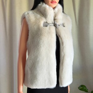 May include: A sleeveless vest made of cream-colored faux fur with a high collar. The garment features a single toggle closure at the upper chest area, consisting of a grey loop and a matching grey toggle fastener to secure the front opening.