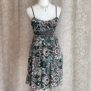 Puede incluir: Vestido sin mangas con estampado de cachemira en negro, verde azulado y blanco. El vestido tiene finas correas negras, cintura con cuentas y falda acampanada. Se ve un collar plateado con dijes de llaves.
