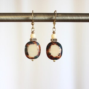 May include: A pair of handcrafted earrings with a cream-colored oval center, surrounded by a dark brown and teal mottled pattern. The earrings have gold-colored hooks, small beads, and decorative accents. The earrings are hanging on a gold-colored rod.