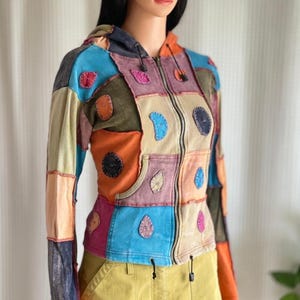 Puede incluir: Una sudadera con capucha y cremallera de patchwork en una variedad de colores, incluyendo azul, naranja, rosa y verde. La chaqueta tiene una capucha, una cremallera frontal y parches decorativos. Las mangas son largas y la chaqueta tiene un cordón en la parte inferior.