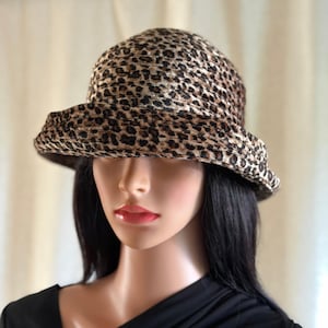 May include: A bucket hat featuring a brown and black leopard print pattern. The hat has a rounded crown and a downward-sloping brim. The fabric has a textured appearance with horizontal stitching lines visible across the crown and the brim.