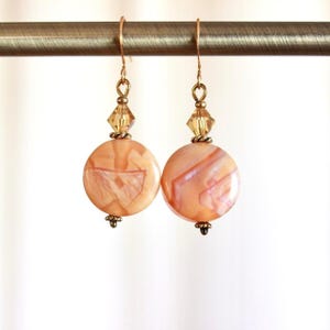 May include: A pair of peach-colored earrings with a round, marbled stone design. The earrings have gold-colored hooks, a small crystal bead, and a decorative bottom piece. The earrings are hanging from a gold-colored bar.