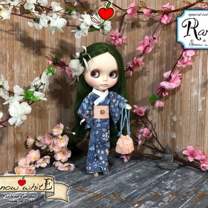 Pode incluir: Um quimono azul e branco estampado com uma faixa rosa e uma bolsa combinando. A boneca está vestindo o quimono e segurando a bolsa. A boneca tem cabelo verde e olhos azuis. O fundo é uma parede de madeira com flores rosas.