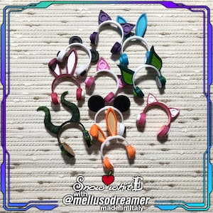 Könnte beinhalten: Eine Sammlung von 12 bunten Kopfhörern im Stirnbandstil mit verschiedenen Tierohr-Designs, darunter Hasen-, Katzen-, Maus- und Drachenohren. Der Text "Snow white with @mellusodreamer made in Italy" ist am unteren Rand des Bildes sichtbar.