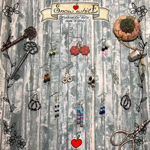 Può includere: Una collezione di 12 paia di orecchini per bambole, con vari design e colori, tra cui nero, bianco, rosso, blu e verde. Gli orecchini sono esposti su uno sfondo di legno con il testo "Snow White Fashion for dolls made in Italy".